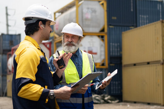 Senior and young male workers in hard hats and safety vests review logistics data on a tablet at a busy container terminal, ideal for industrial and supply chain content.