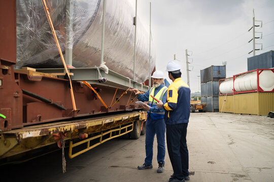 Two logistics workers inspecting heavy cargo on flatbed trailer at industrial shipping yard with containers