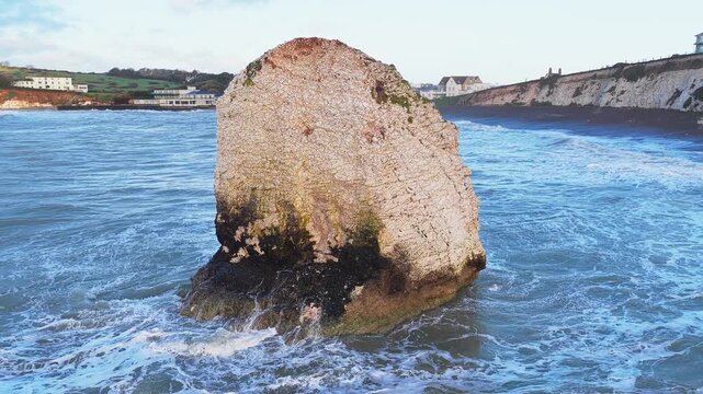 Aerial drone orbit around Stag Rock at Freshwater Bay, Isle of Wight, showing waves crashing against cliffs and rocky shoreline on a cloudy winter day.