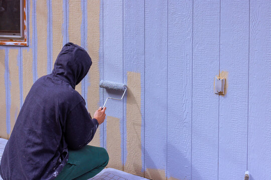 Painter uses roller to paint siding wall blue while crouching down outside house