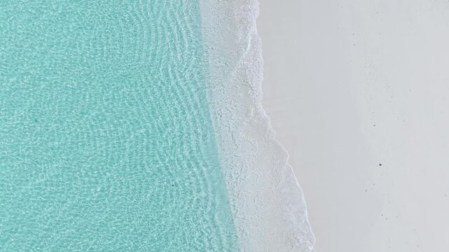Aerial view of crystal clear turquoise water gently lapping against a pristine white sand beach with soft ripples and bright sunlight in Vashafaru, Haa Alif Atoll, Maldives.