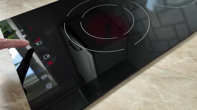 A close-up of a modern induction cooktop with a hand pressing the control panel. The surface is sleek and black, reflecting light.