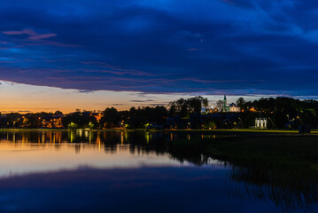 Naklejka premium Beautiful summer evening night view of a small town Telsiai, Lithuania by the lake, night lighting lamps reflected in the lake, dramatic blue sky with clouds, urban landscape, copy space.Long exposure