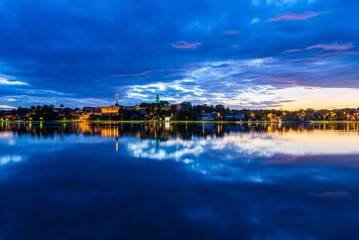 Fototapeta premium Beautiful summer evening night view of a small town Telsiai, Lithuania by the lake, night lighting lamps reflected in the lake, dramatic blue sky with clouds, urban landscape, copy space.Long exposure