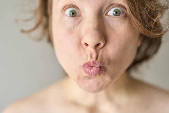 Woman Puckering Lips With Wide Open Eyes Making Playful Surprised Face In Neutral Studio Setting Closeup Portrait Bare Shoulders Under Bright Natural Light.