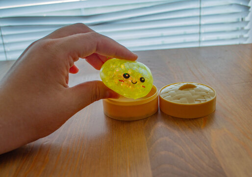 Close-up: a person's hand reaches for a yellow shiny squishy with a cute face over an open steamer container. The lid lies nearby on the polished wooden table.