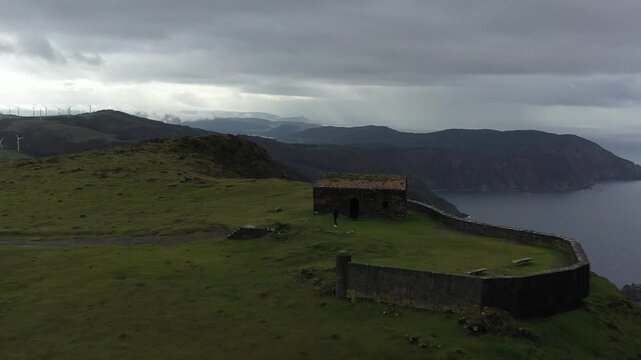 Aerial view of Garita de Herbeira stone structure on a cliff overlooking the sea with green hills and wind turbines under an overcast sky in Meizoso, A Coruna, Spain.