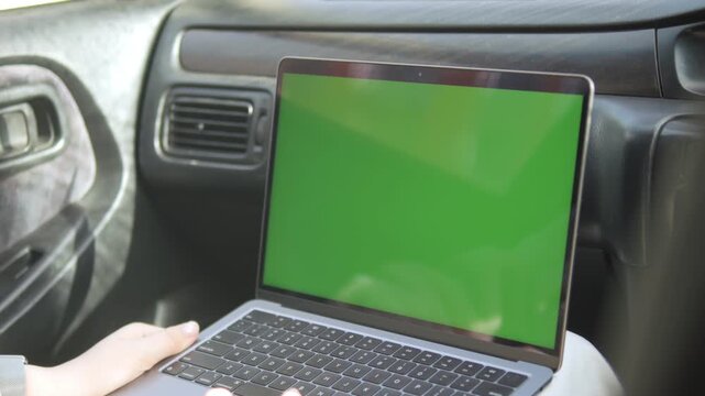 Person uses a laptop with green screen inside a car. Hands rest on keyboard, focused and calm. This footage suits travel, remote work, or car-based digital projects. No emotion visible