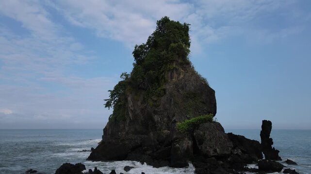 Aerial view of Pantai Karang Agung Kebumen, a massive rock formation covered in lush greenery rising from the crashing waves of the Indian Ocean Ayah, Central Java, Indonesia.