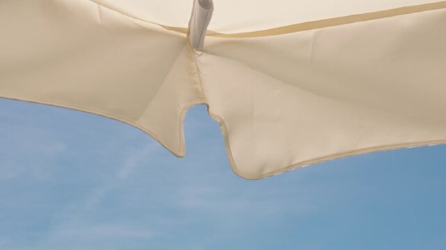 Looking up at patio umbrella moving in wind on sunny summer day. POV unique low angle view from underneath a large cream garden umbrella gently swaying in the breeze against a clear blue sky. 