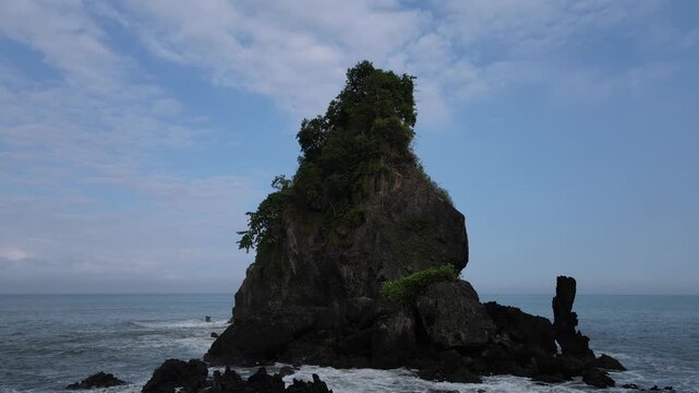 Aerial view of Pantai Karang Agung Kebumen sea stack with green trees on top and waves crashing against the dark rocks under a blue sky in Ayah, Central Java, Indonesia.