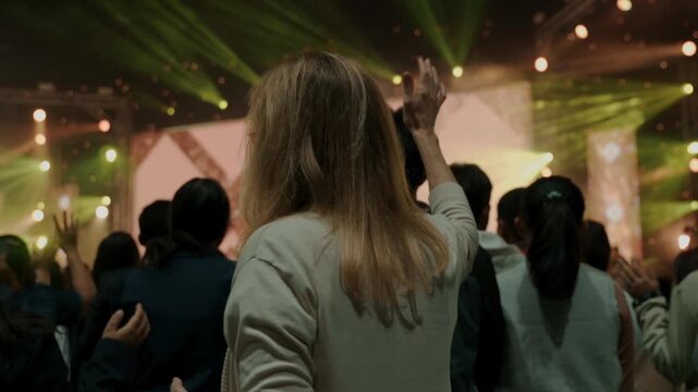 Rear view of a woman raising her hand during a worship concert surrounded by a crowd. Spiritual atmosphere, faith, praise and worship, religious gathering, community celebration, and inspirational mus