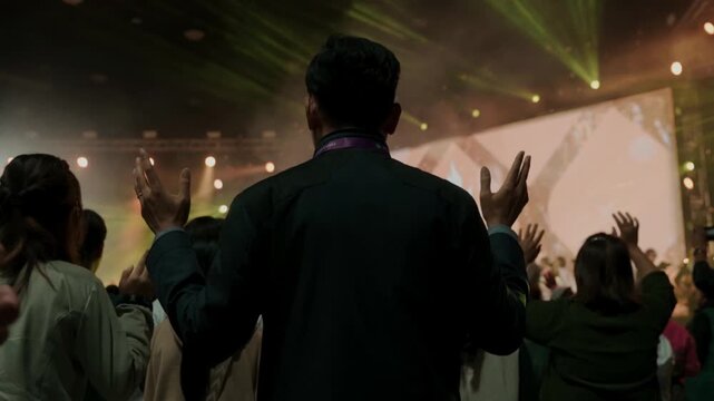 Rear view of a man standing among a crowd during a live worship concert. People raising hands in front of a brightly lit stage, expressing faith, hope, spirituality, community, and emotional connectio