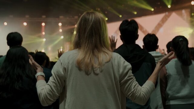 Rear view of a woman raising her hand during a worship concert surrounded by a crowd. Spiritual atmosphere, faith, praise and worship, religious gathering, community celebration, and inspirational mus