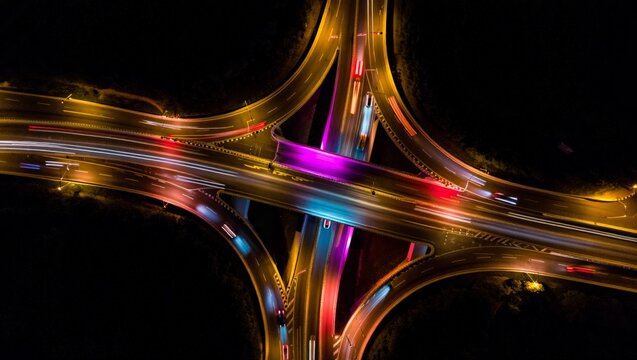 Stunning drone aerial of a major urban road junction at night with spectacular multicolor light trails in orange, blue, pink and white weaving through overpasses and curved ramps on a black background