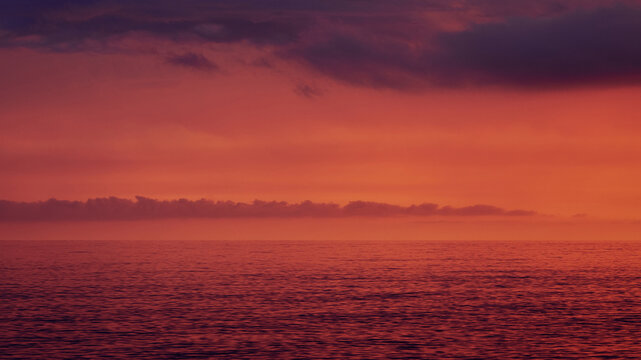 A breathtaking minimalist seascape at sunset in Scalea, Italy. The dramatic red and purple sky is filled with dark clouds, beautifully reflecting over the calm waters of the Mediterranean Sea.