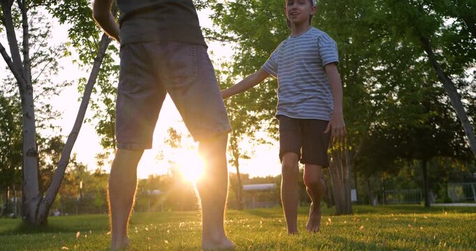 Father and son play soccer barefoot in park at sunset as boy celebrates successful kick with raised hands. Horizontal outdoor scene captures joyful family bonding and active parenting