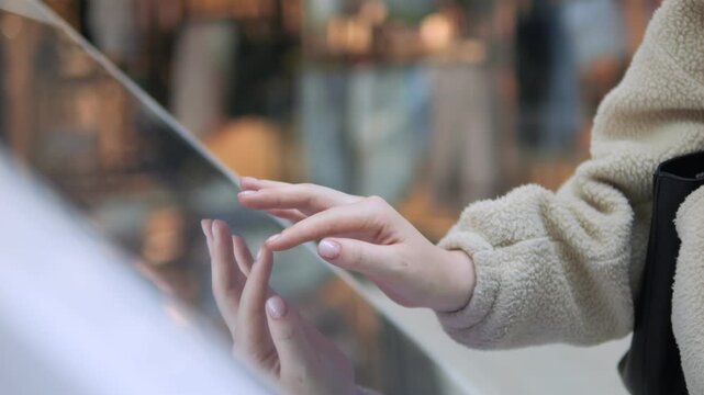 Person taps glass surface with fingers. Hands shown in close-up detail. Background blurred, urban setting visible. Focus on touch interaction with reflective surface