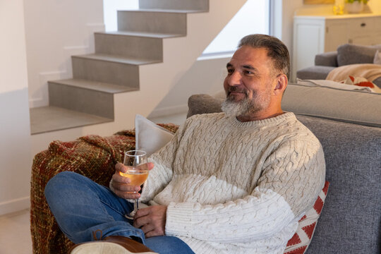 Senior man sitting on gray sofa in living room holding stemmed wine glass wearing cream sweater