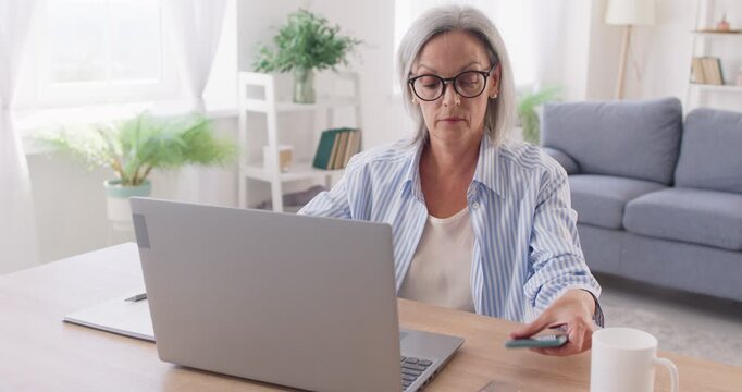 Mature woman takes notes while working on laptop and is distracted by her smartphone. Woman looks at screen of her mobile phone while working, smiling at something that catches her interest. 4K video.