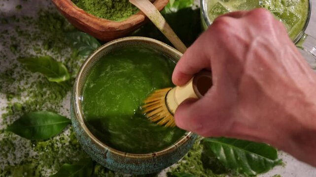 Hand whisking vibrant green matcha tea with traditional bamboo whisk, surrounded by matcha powder and tea-making ingredients. Illuminated by natural sunlight, creating an authentic tea ceremony scene