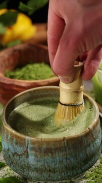Hand whisking vibrant green matcha tea with traditional bamboo whisk, surrounded by matcha powder and tea-making ingredients. Illuminated by natural sunlight, creating an authentic tea ceremony scene
