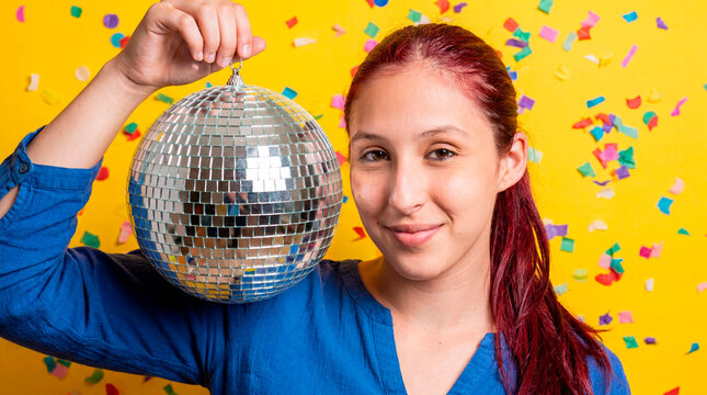 Confident latina woman holding disco mirror ball on yellow background with falling confetti