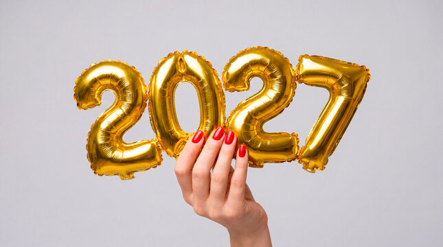 Womans manicured hand holding golden 2027 foil balloons on minimal gray background