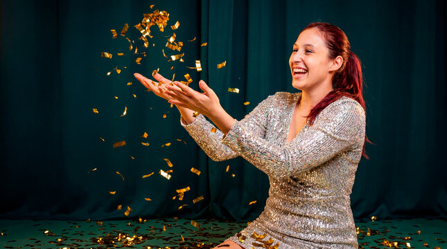 Joyful latina woman in sequin dress throwing golden confetti against green velvet curtain