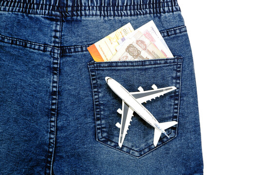 A white airplane is placed in the back pocket of blue jeans pant with folded foreign banknotes peeking out of the top of the pocket, suggesting travel and expenses.