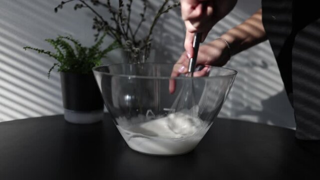 Female hands whisk egg whites in a transparent glass bowl creating a smooth airy foam Close up baking process with focus on texture movement and homemade cooking