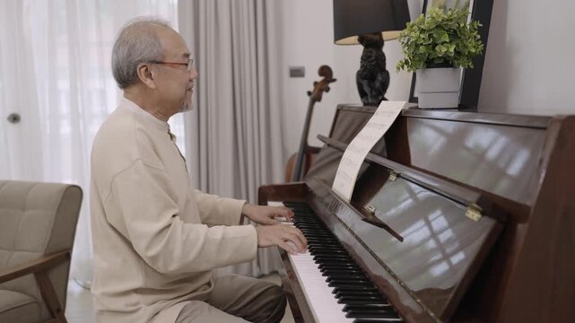 Asian senior man pianist in eyeglasses is sitting playing classical piano alone after retirement for relaxing in living room at home. Elderly male playing a keyboard organ instrument with happiness. 