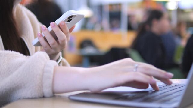 Person uses a phone while typing on a laptop. Hands hold the phone and keyboard. Background shows blurred people. Focus remains on hands and devices. Scene suggests multitasking or communication