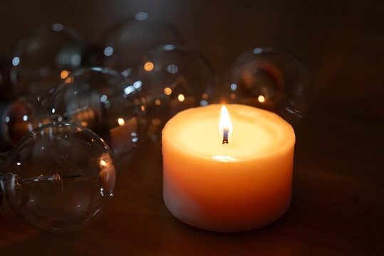 A lit wax candle burning next to glass light bulbs in the dark. A candle illuminates turned-off lamps symbolizing resilience alternative energy and hope during a blackout