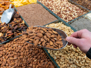 Obraz premium A metal scoop with almonds in a customer's hand over a store counter. The process of choosing fresh nuts at the market symbolizing quality food traditions and a healthy lifestyle