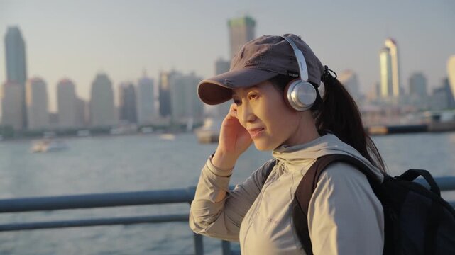 A young Asian female backpacker wearing a cap and headphones stands by a riverside railing, enjoying the urban waterfront city skyline at sunset.