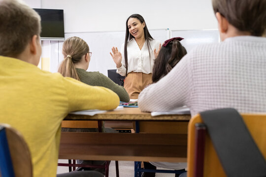 A small group of students is in their elementary classroom while a mixed-race teacher guides them through a math and geometry lesson. She is asking questions and helping them stay focused.