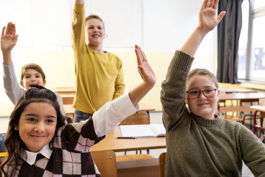 The teacher asks a question, and several students eagerly raise their hands to answer. The moment feels active and positive, with everyone engaged in the lesson.
