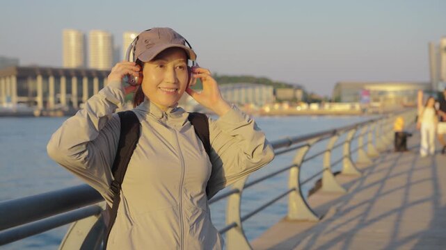 A young female athlete adjusts her headphones while standing by a waterfront city promenade during golden hour, enjoying post-workout relaxation.
