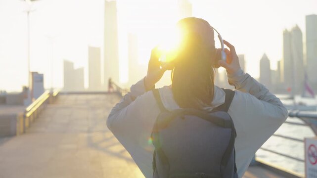 A rear view of a female backpacker walking along a waterfront promenade at sunset, overlooking a downtown city skyline under warm golden hour light.