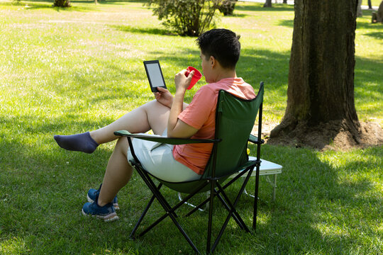 A young person drinking and reading an e-book in a sunny park