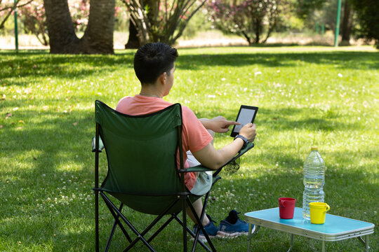 A young Caucasian person reading an e-book while relaxing in the park