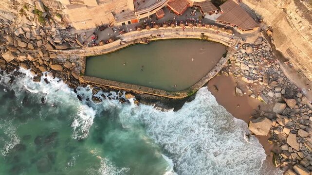 Aerial view of Azenhas do Mar natural swimming pool with waves crashing against the cliffs and people relaxing by the water in Colares, Lisbon, Portugal.
