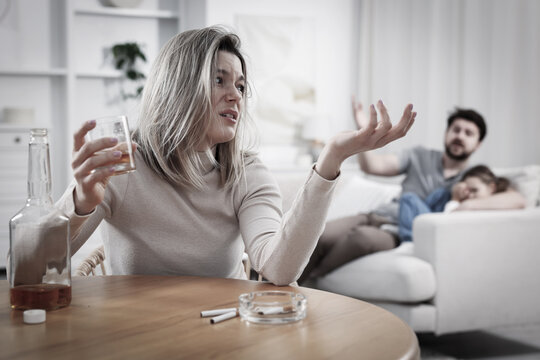 Alcohol addiction. Woman drinking whiskey at wooden table while her unhappy husband and daughter sitting on sofa indoors, selective focus