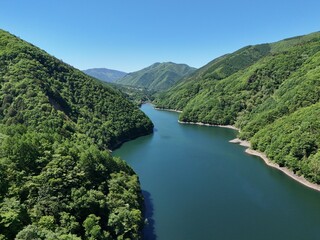 Fototapeta premium Lush green hills embrace calm, deep blue river. Clear sky above adds serene, natural beauty. Perfect for tranquil nature scenes or travel inspiration. No animals or people visible in the image