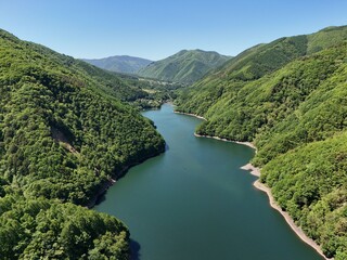 Fototapeta premium Lush green hills frame a calm, winding river. Deep blue waters reflect the clear sky above. Rugged slopes covered in dense foliage rise gently on either side. The scene radiates peace