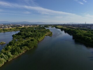 Fototapeta premium Aerial view of a wide river flowing through lush greenery. Distant mountains frame the horizon under a clear blue sky. Vibrant trees line both riverbanks, adding natural depth and contrast