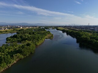 Fototapeta premium Serene river winds through lush greenery. Blue sky stretches above with wispy clouds. Flooded banks frame calm, reflective water. Distant cityscape blends with nature's edge