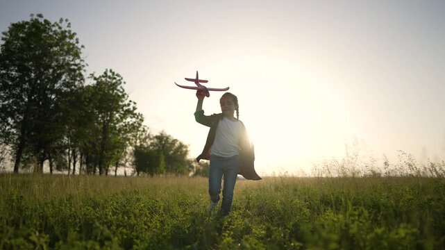 Girl runs through field at sunset holding red toy airplane above head. Child plays with plane in tall grass. Kid dreams of flight. Happy girl enjoys evening play outdoor. Young pilot imagines flying.