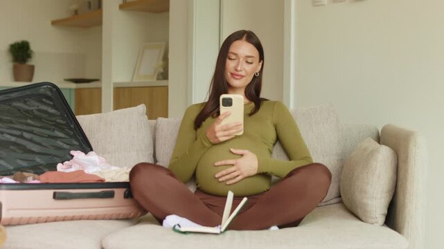 Pregnant woman using smartphone on sofa at home and tenderly touching belly